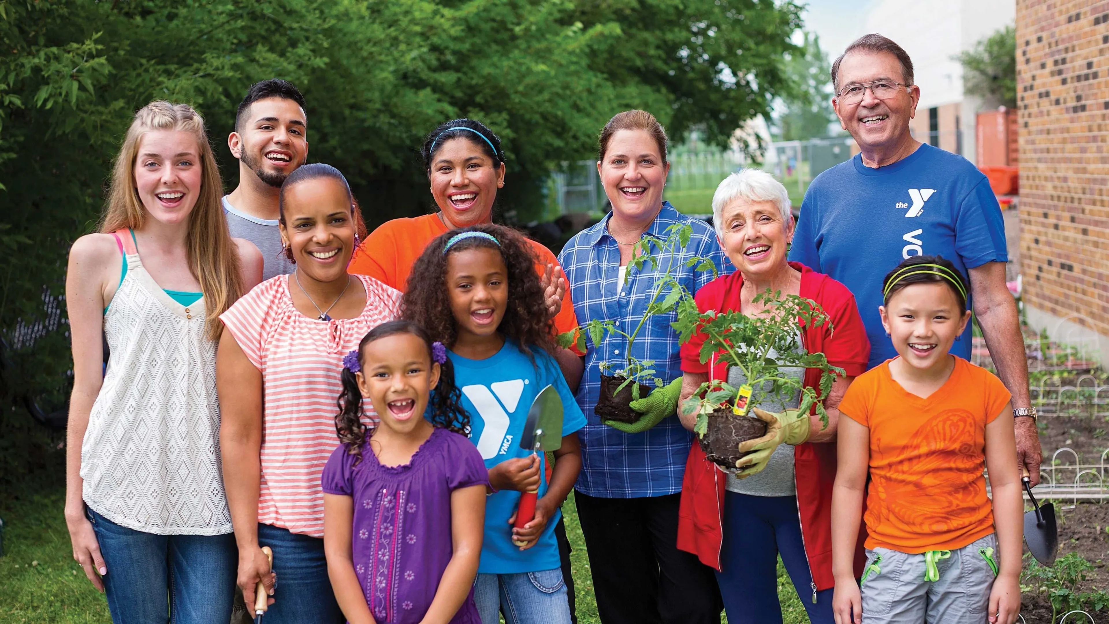 Group of diverse people smile, posing for a photo together in the garden. Kids and adults hold plants, gardening tools; a community activity.