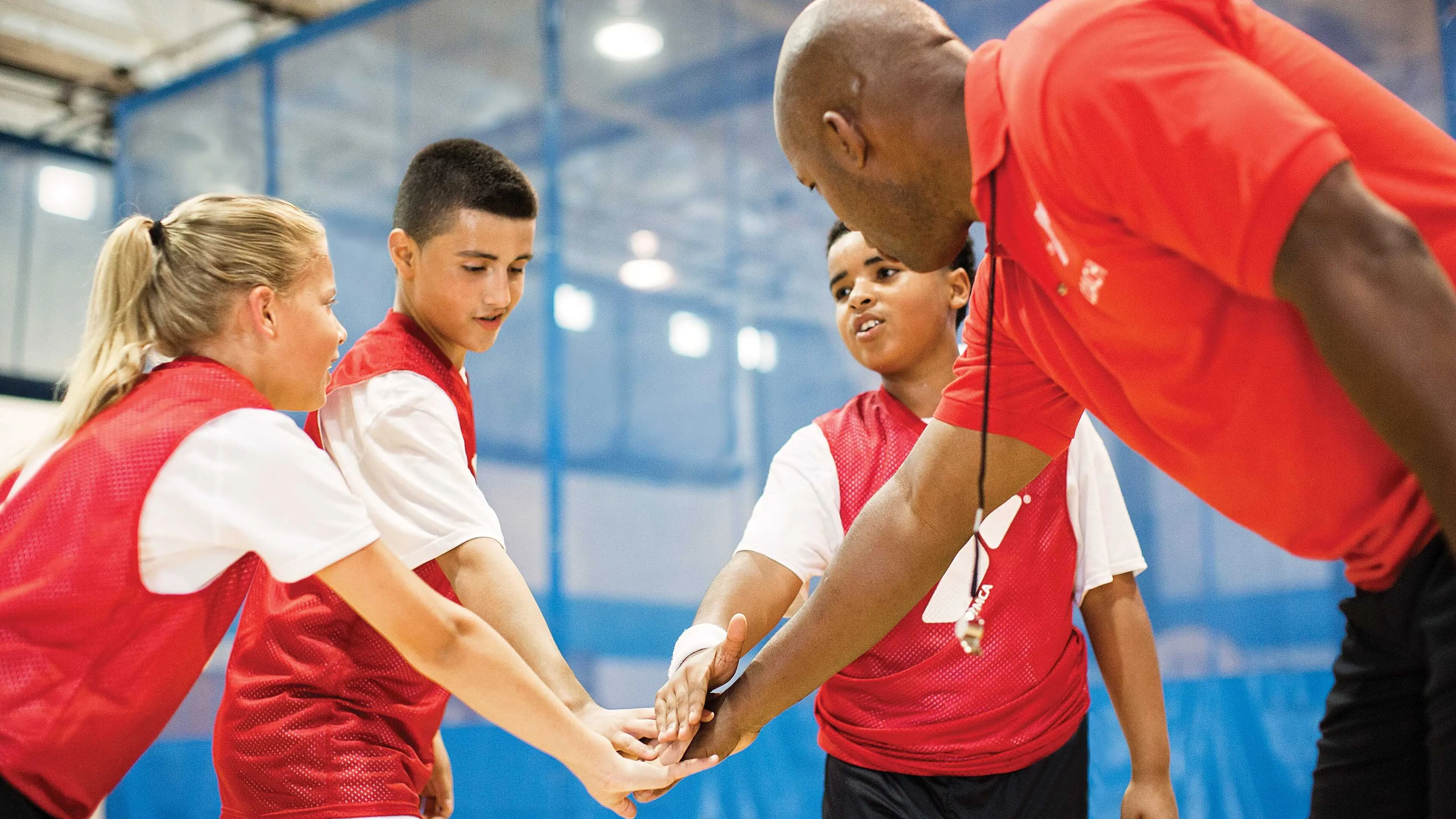 A coach and three kids in red vests with hands stacked in the middle in a team spirit gesture, indoors.