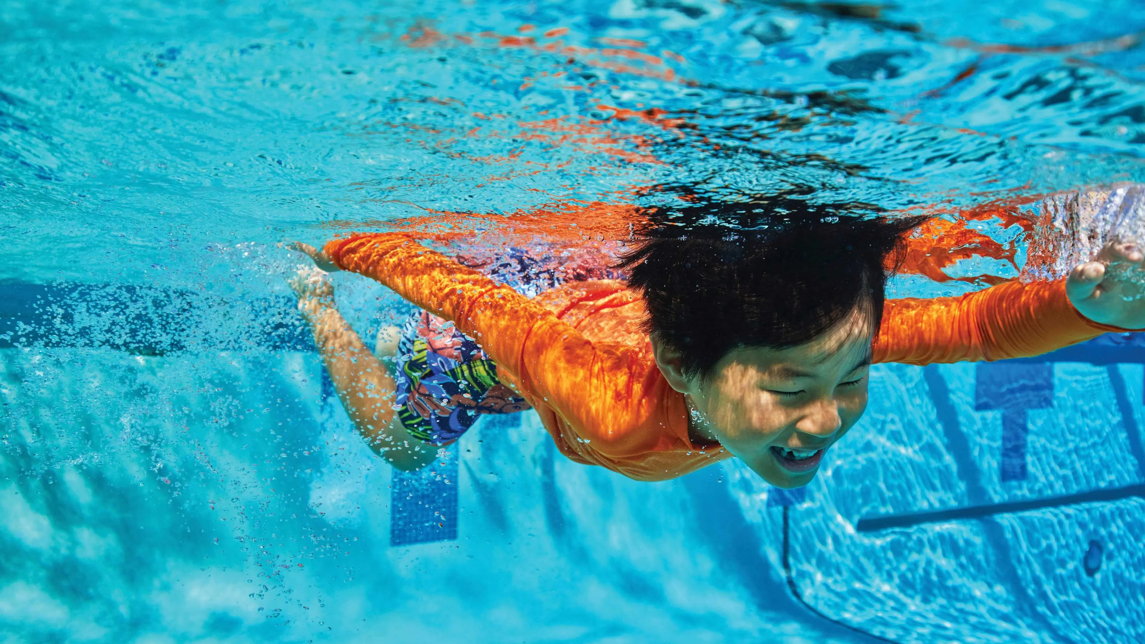 Young boy in an orange shirt swims underwater in a pool, arms stretched forward and smiling. Clear blue water surrounds him.