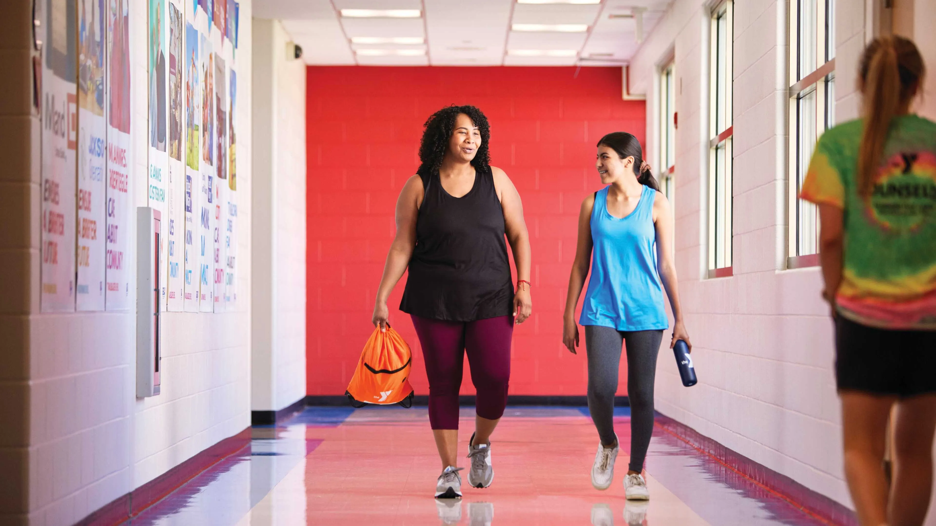 Two women are walking and talking in a brightly lit hallway with a third person walking away. One woman carries a bag, another a water bottle.