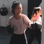 Two women in boxing gloves practice in a gym setting, one smiling with a punch; fitness program for all ages.

