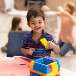 A delighted young boy holds a yellow block, smiling widely near a building block structure, and plays with others at a kid's program.
