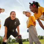 A smiling coach and three kids in baseball attire huddle and share a laugh on a sunny day during a sports program.
