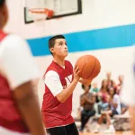 Teenager in a red jersey about to shoot a basketball during a game, surrounded by teammates and spectators in the background.
